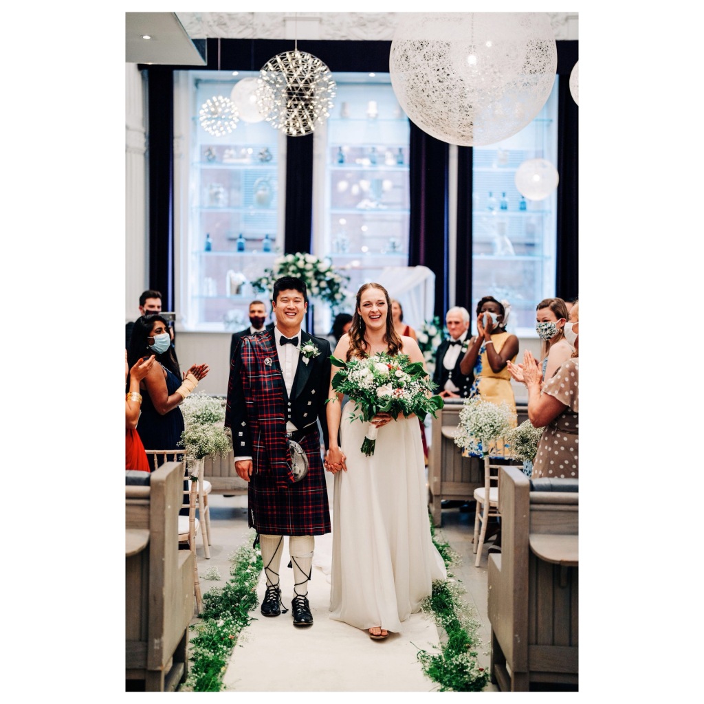 A different sex couple walk down their aisle holding hands and smiling, their guests stand and clap. The guests are wearing facemasks. The groom is in traditional kilt attire and the bride in a long white dress holds a large bouquet.