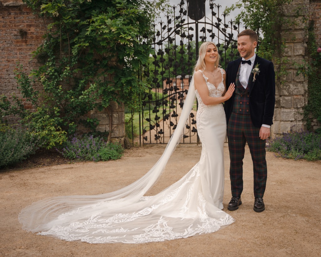 A white, different sex couple stand close. She is in a long white, spaghetti strap dress with long train and veil. He is in tartan trousers and waistcoat with a velvet jacket. They are standing at wrought iron gates both smiling