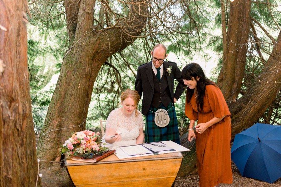 A woman in white sits at a table covered on flowers and folders and paperwork. She holds a pen. A man in traditional kilt attire stands behind her and looks down smiling. The celebrant hovers close by. They are amongst the spreading trunks and branches of a magical tree.